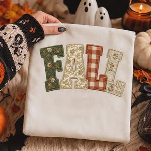 May include: A white sweatshirt featuring the word FALL in large, block-style letters. Each letter has a unique fabric pattern: the F is dark green with floral quilting, the A is cream with floral print, the L is brown gingham, and the L is plaid.