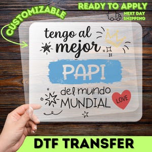 Könnte beinhalten: Eine transparente DTF-Transferfolie mit dem schwarzen Text tengo al mejor PAPI del mundo MUNDIAL, einem blauen Pinselstrich-Hintergrund hinter dem Wort PAPI, einer gelben Krone, schwarzen Sternsymbolen und einem roten Herz mit dem Wort LOVE.
