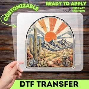 May include: A clear DTF transfer sheet featuring a desert landscape illustration with a tall saguaro cactus, smaller cacti, and mountains under a sun with radiating orange and cream rays. Three birds fly in the sky above the mountain range.