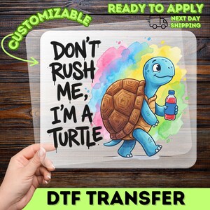 May include: A clear DTF transfer sheet featuring a cartoon blue turtle with a brown shell holding a red water bottle. The text reads DON'T RUSH ME, I'M A TURTLE in black graffiti-style lettering against a colorful watercolor splash background.