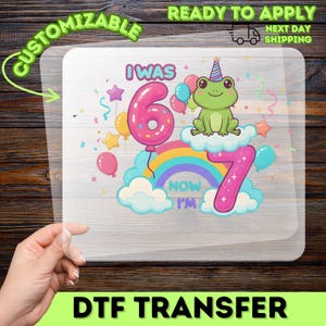 May include: A DTF transfer featuring a colorful design for a 7th birthday. The design includes a large pink number 6 balloon, a rainbow, a frog wearing a party hat, and the text "I WAS 6, NOW I'M 7".