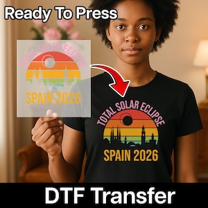 Op de afbeelding: Zwart T-shirt met een kleurrijk ontwerp van een zonsondergang, een gedeeltelijke zonsverduistering en de tekst "SPAIN 2026". De afbeelding toont ook een DTF-transfer met hetzelfde ontwerp en de woorden "Ready To Press".