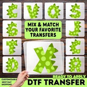 May include: A collection of green and white DTF transfers featuring letters and numbers with a dinosaur-like design. The image includes the text "MIX & MATCH YOUR FAVORITE TRANSFERS" and "READY TO APPLY DTF TRANSFER".