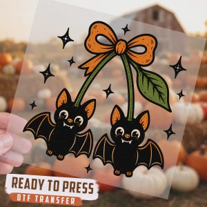 May include: A DTF transfer sheet featuring a graphic of two black bats hanging like cherries from a single orange bow with green stems and a leaf. The design includes small black star accents and the text READY TO PRESS DTF TRANSFER at the bottom.