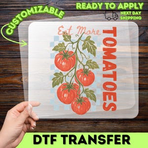 Op de afbeelding: Een transparant DTF-transfervel met een afbeelding van een wijnstok met vier rode tomaten en groene bladeren. De tekst luidt Eat More TOMATOES in rood en lichtoranje. De achtergrond van de afbeelding heeft een lichtblauw en wit ruitpatroon.