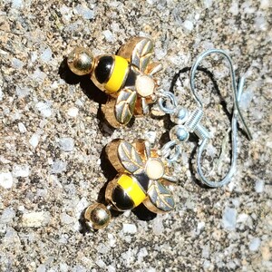 Puede incluir: Un par de pendientes de abeja dorados con detalles de esmalte negro y amarillo. Los pendientes tienen una pequeña bola dorada en la parte superior y están sujetos a ganchos plateados. Las alas de la abeja están detalladas con líneas grabadas.