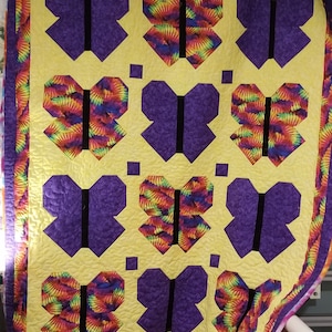 May include: A handmade quilt featuring a repeating butterfly pattern. The butterflies alternate between solid purple and a vibrant tie-dye design in yellow, orange, green, and blue. The quilt has a yellow background and a purple border.