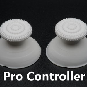 Peut inclure: Deux remplacements de joystick blancs texturés en forme de dôme pour une manette de jeu. Le texte "Pro Controller" est imprimé sous les joysticks.