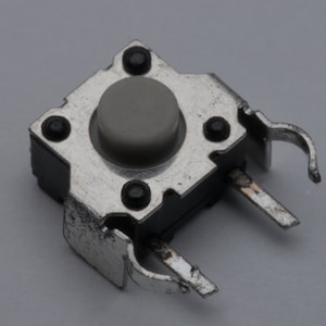 May include: A small, square electronic component featuring a grey button and a silver metal frame. The component has four black mounting pins and two silver metal tabs extending from the sides. The button is slightly raised and centered on the component.