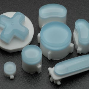 May include: A collection of replacement gaming controller buttons featuring a white base and a translucent light blue top. The set includes a directional pad, various circular buttons of different sizes, and a long rectangular button.