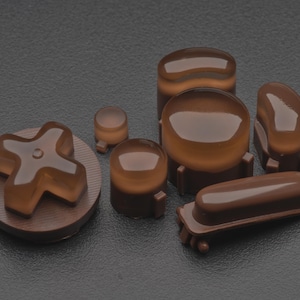 Peut inclure: Ensemble de boutons de manette de jeu bruns, comprenant une croix directionnelle, des boutons ronds et un bouton rectangulaire. Les boutons ont un aspect translucide et sont disposés sur une surface sombre. Accessoires ou pièces de rechange.