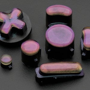 Op de afbeelding: Een set van acht iriserende paarse en zwarte controllerknoppen voor een gameconsole. De knoppen zijn gemaakt van plastic en hebben een glanzende afwerking.