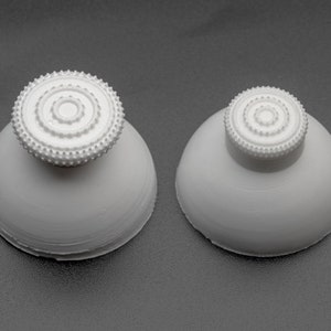 Może przedstawiać: Dwa białe plastikowe przyciski do joysticka w kształcie kopuły z fakturowanym okrągłym wzorem na górze.