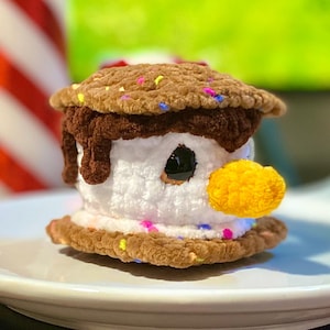 May include: A crocheted dessert resembling a s'more with a snowman face. The s'more has a brown cookie base and top, with a white marshmallow center. The snowman has a yellow beak and black eyes, and is decorated with colorful sprinkles.