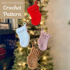 May include: Four mini crocheted stockings in red, white, brown, and pink hanging on a Christmas tree. The stockings have a pom-pom trim and are decorated with beads. The image includes the text "Mini Stockings Crochet Pattern".