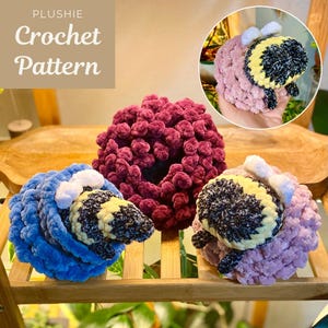 May include: Three crocheted bee plushies, one blue, one pink, and one burgundy. The plushies are sitting on a wooden shelf with green plants in the background. The image includes the text "PLUSHIE Crochet Pattern".