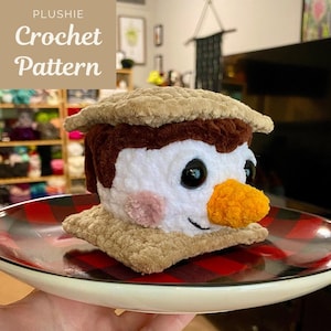 May include: A crocheted plush toy shaped like a s'more. The toy has a brown chocolate layer, a white marshmallow layer, and a tan graham cracker layer. The marshmallow layer has a smiling face with black eyes and a yellow nose.