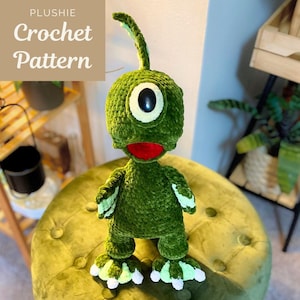 May include: A green crocheted plushie with a large eye, red mouth, and a curved antenna. The plushie is standing on a green velvet ottoman. The text "PLUSHIE Crochet Pattern" is in the upper left corner.