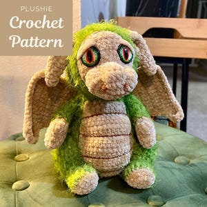 May include: A crocheted dragon plushie in green and beige tones. The dragon features large, detailed eyes and wings. The text "PLUSHIE Crochet Pattern" is visible in the upper left corner. The dragon is sitting on a green surface.