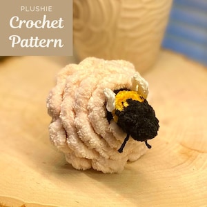 May include: A crocheted bee plush toy with black and yellow stripes, white wings, and black eyes. The bee is peeking out of a pink crocheted nest.