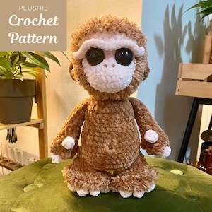 May include: A handmade brown crochet monkey plushie with a white face, large black eyes, and white accents on its hands and feet. The text "PLUSHIE Crochet Pattern" is in the upper left corner. The plushie is sitting on a green surface.