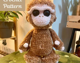 May include: A handmade brown crochet monkey plushie with a white face, large black eyes, and white accents on its hands and feet. The text "PLUSHIE Crochet Pattern" is in the upper left corner. The plushie is sitting on a green surface.