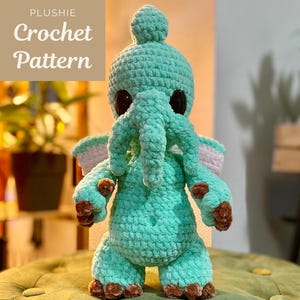 Op de afbeelding: Een turquoise gehaakte olifantenknuffel met grote zwarte ogen, bruine voeten en roze en witte accenten op de oren. De knuffel staat op een groen oppervlak. De tekst "PLUSHIE Crochet Pattern" staat in de linkerbovenhoek.