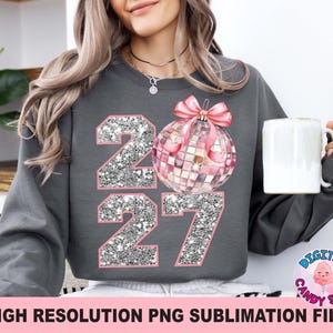 May include: A charcoal gray long-sleeve sweatshirt featuring a graphic of the number 2027 with a silver glitter texture and pink outlines. A pink disco ball ornament with a pink bow is positioned over the zero, replacing the digit.