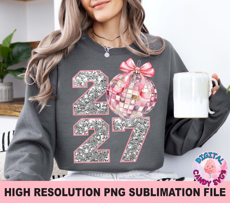 May include: A charcoal gray long-sleeve sweatshirt featuring a graphic of the number 2027 with a silver glitter texture and pink outlines. A pink disco ball ornament with a pink bow is positioned over the zero, replacing the digit.