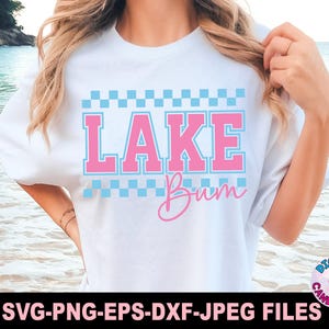 May include: White t-shirt with the text "LAKE Bum" in pink and blue. The word "LAKE" is in a block letter style with a checkered pattern above and below. The word "Bum" is in a cursive font.