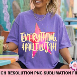 May include: A purple t-shirt with the words "EVERYTHING, HALLELUJAH" in yellow script over a pink star. The shirt is a relaxed fit and has a vintage look. The image also includes a drink in a clear cup.