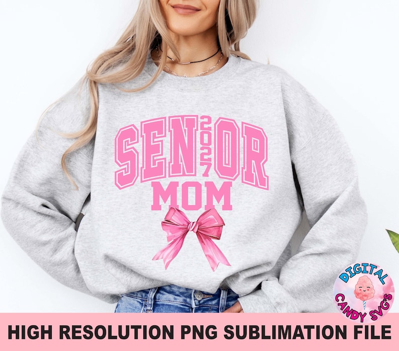 Pode incluir: Uma camisola de gola redonda cinzenta clara com o texto cor-de-rosa SENIOR 2027 MOM numa fonte universit&aacute;ria. Por baixo do texto encontra-se um grande la&ccedil;o de fita cor-de-rosa ilustrado. O design est&aacute; centrado no peito da pe&ccedil;a.