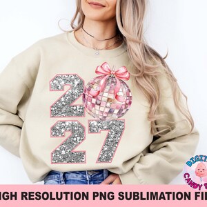 May include: A beige crewneck sweatshirt featuring a graphic of the year 2027 in silver glitter with pink outlines. A pink disco ball ornament with a pink bow is positioned between the numbers, replacing the zero in the year 2027 design.