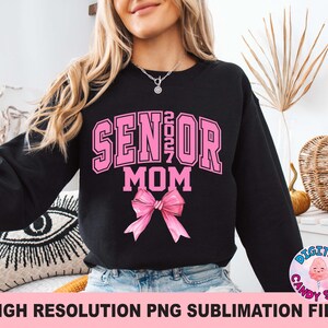 Pode incluir: Uma camisola preta de manga comprida com o texto cor-de-rosa SENIOR 2027 MOM numa fonte universit&aacute;ria. Abaixo do texto encontra-se um gr&aacute;fico de um la&ccedil;o cor-de-rosa. O design est&aacute; centrado no peito da pe&ccedil;a, usada sobre cal&ccedil;as de ganga azuis.