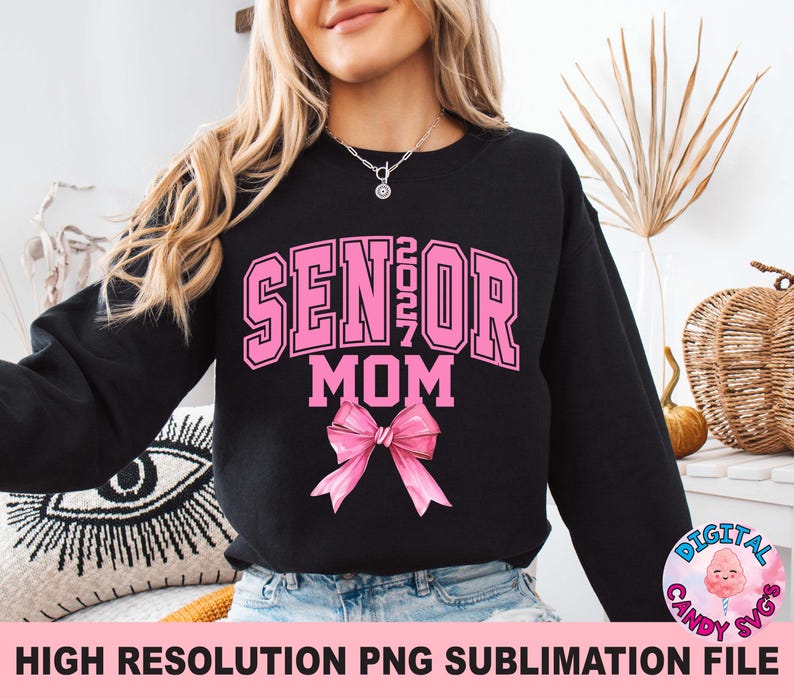 Pode incluir: Uma camisola preta de manga comprida com o texto cor-de-rosa SENIOR 2027 MOM numa fonte universit&aacute;ria. Abaixo do texto encontra-se um gr&aacute;fico de um la&ccedil;o cor-de-rosa. O design est&aacute; centrado no peito da pe&ccedil;a, usada sobre cal&ccedil;as de ganga azuis.