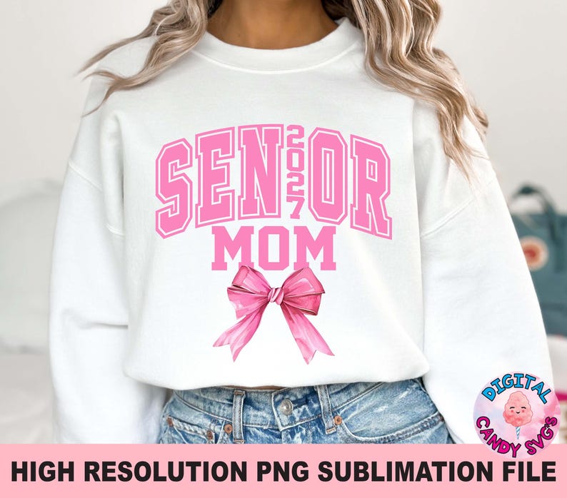 Pode incluir: Uma camisola branca de manga comprida com o texto rosa SENIOR 2027 MOM numa fonte universit&aacute;ria. Abaixo do texto encontra-se um grande la&ccedil;o rosa ilustrado com sombreado em estilo aguarela. O design est&aacute; centrado na frente da pe&ccedil;a.