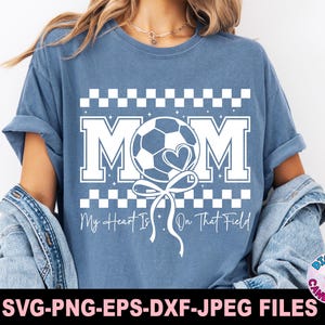 May include: A blue t-shirt with a white checkered pattern and the word "MOM" in white letters. A white soccer ball with a heart is attached to a white ribbon and is positioned between the "M" and the "O". The text "My Heart Is On That Field" is below the design.