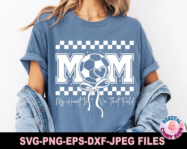 May include: A blue t-shirt with a white checkered pattern and the word "MOM" in white letters. A white soccer ball with a heart is attached to a white ribbon and is positioned between the "M" and the "O". The text "My Heart Is On That Field" is below the design.