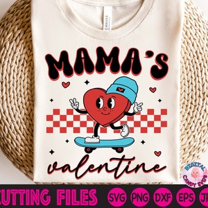 Op de afbeelding: Een wit T-shirt met de tekst "Mama's Valentine" in zwart en rood. Een cartoonhart met een blauwe hoed skateboarded op een geruit patroon.