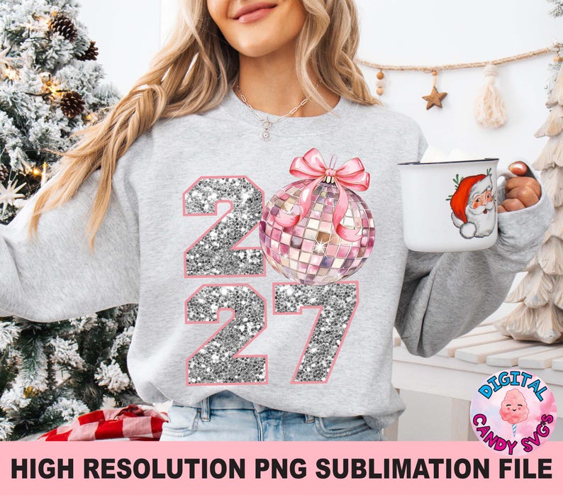May include: A light gray crewneck sweatshirt featuring a graphic of the year 2027 in silver glitter texture, with a pink disco ball ornament replacing the zero. The sweatshirt is paired with a white mug displaying a small illustration of Santa Claus.