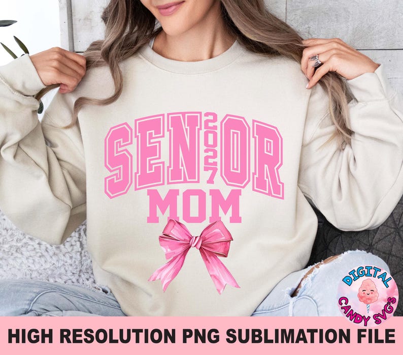 Pode incluir: Uma camisola bege de gola redonda com o texto rosa SENIOR 2027 MOM numa fonte universit&aacute;ria. Abaixo do texto encontra-se um gr&aacute;fico de um la&ccedil;o rosa grande e detalhado. O design est&aacute; centrado no peito da pe&ccedil;a de manga comprida.