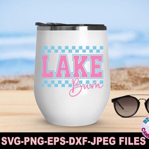 May include: A white, stemless wine tumbler with a black lid, featuring the text "LAKE Bum" in pink and blue. The design includes a checkered border. Sunglasses rest on a wooden surface, with a beach backdrop.