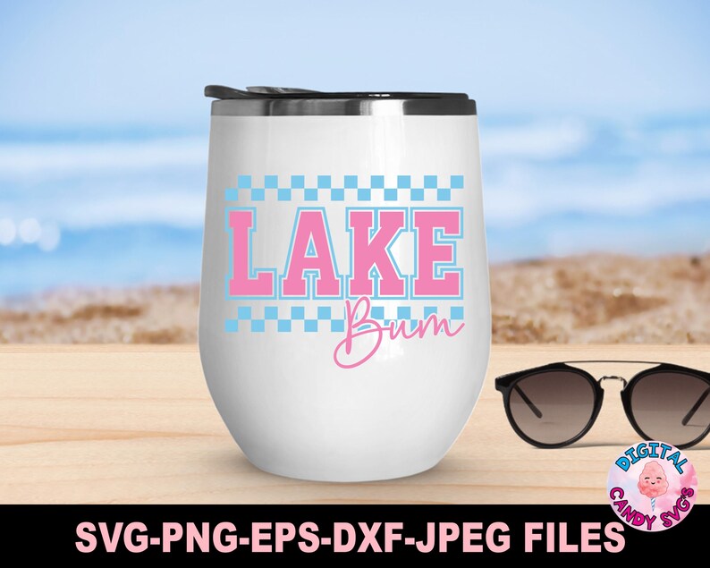 May include: A white, stemless wine tumbler with a black lid, featuring the text "LAKE Bum" in pink and blue. The design includes a checkered border. Sunglasses rest on a wooden surface, with a beach backdrop.