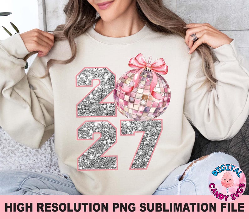 May include: A beige crewneck sweatshirt featuring a graphic print of the number 2027 with a silver glitter texture and a pink disco ball ornament with a matching pink bow. The design is centered on the front of the garment.