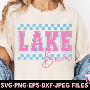 May include: Cream-colored t-shirt with the words "LAKE Bum" in pink and blue. The word "LAKE" is in a bold, collegiate-style font, with a blue checkered pattern above and below. The word "Bum" is in a cursive script.