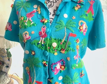 May include: A turquoise blue knit cardigan with a golf theme. The short-sleeved sweater features embroidered golfers, palm trees, flowers, and golf balls. The cardigan has a collared neckline and button-up front. The bottom half of the mannequin is wearing a green skirt.