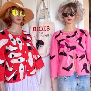May include: Two mannequins wearing cardigans. One is red with white and black shoe designs, the other is pink with black and pink shoe designs. A tote bag with the text "BOOKS not BOMBS" is visible.