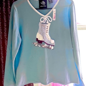 May include: A light blue long-sleeve sweater with a V-neckline and white trim. The front features an appliqué of a white ice skate with silver sequins, white laces, and a pom-pom detail. The interior label displays the text Michael Simon.