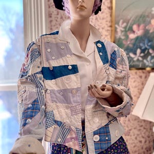 May include: A patchwork jacket with a white collar and buttons. The jacket is made of various shades of blue, white, and purple fabric. The jacket is worn by a mannequin with purple hair and a black hat.
