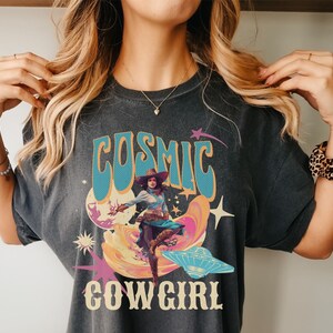 May include: A dark grey t-shirt with a retro graphic design featuring a woman in a cowboy hat riding a cosmic wave. The text "COSMIC COWGIRL" is printed on the shirt.